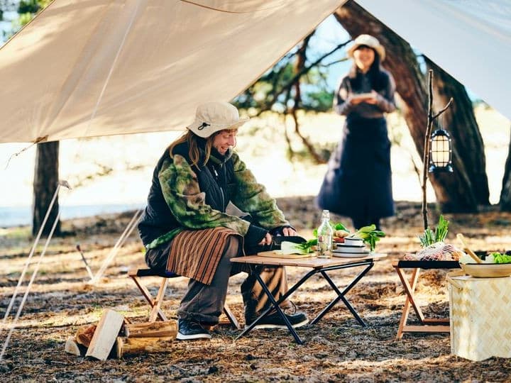 .En person sitter under ett tält vid ett campingbord med mat. En annan person står i bakgrunden.