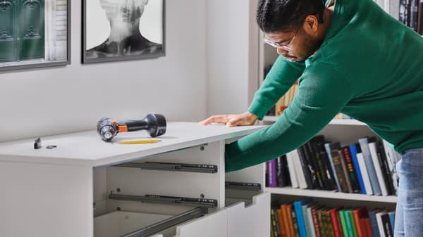 En person strekker armen inn i en hvit NORDLI kommode med sju skuffer der de øverste frontene ennå ikke er påmontert og med en FIXA skrutrekker oppå.