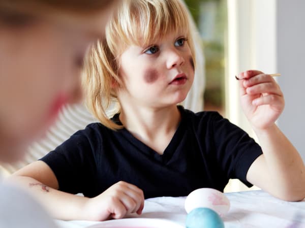 En pojke i svart t-shirt med målade kinder som en påskkärring sitter vid ett bord och målar ägg. 
