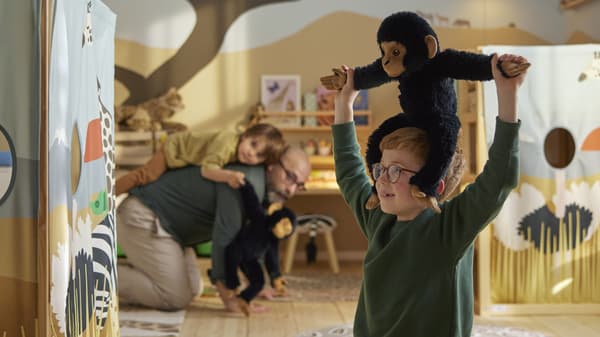 En pojke med en mjukdjursschimpans på axlarna och en vuxen person med ett barn på ryggen leker i ett rum.
