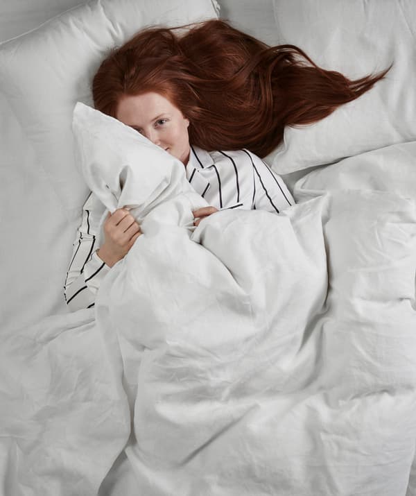 En pyjama rayé, allongée sur le dos dans un lit double, une femme aux longs cheveux roux tient une couette entre les mains.