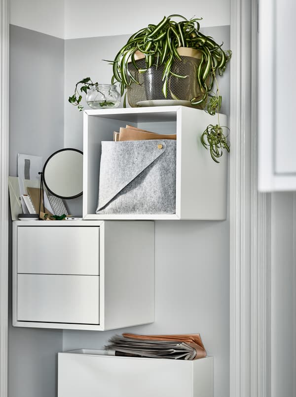 En smal væg ved siden af en dør med hvide, vægmonterede EKET skabe. Et skab er åbent med en plante ovenpå, det andet er med skuffer.