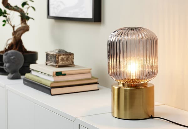 En SOLKLINT lampe med gråt glas og messingfod står ved siden af en stak bøger.