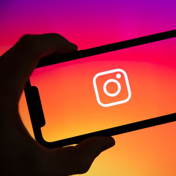 En sort silhuet af en hånd ses holde en sort silhuet af en telefon med Instagram logo i hvidt på en baggrund af Instagrams farver  