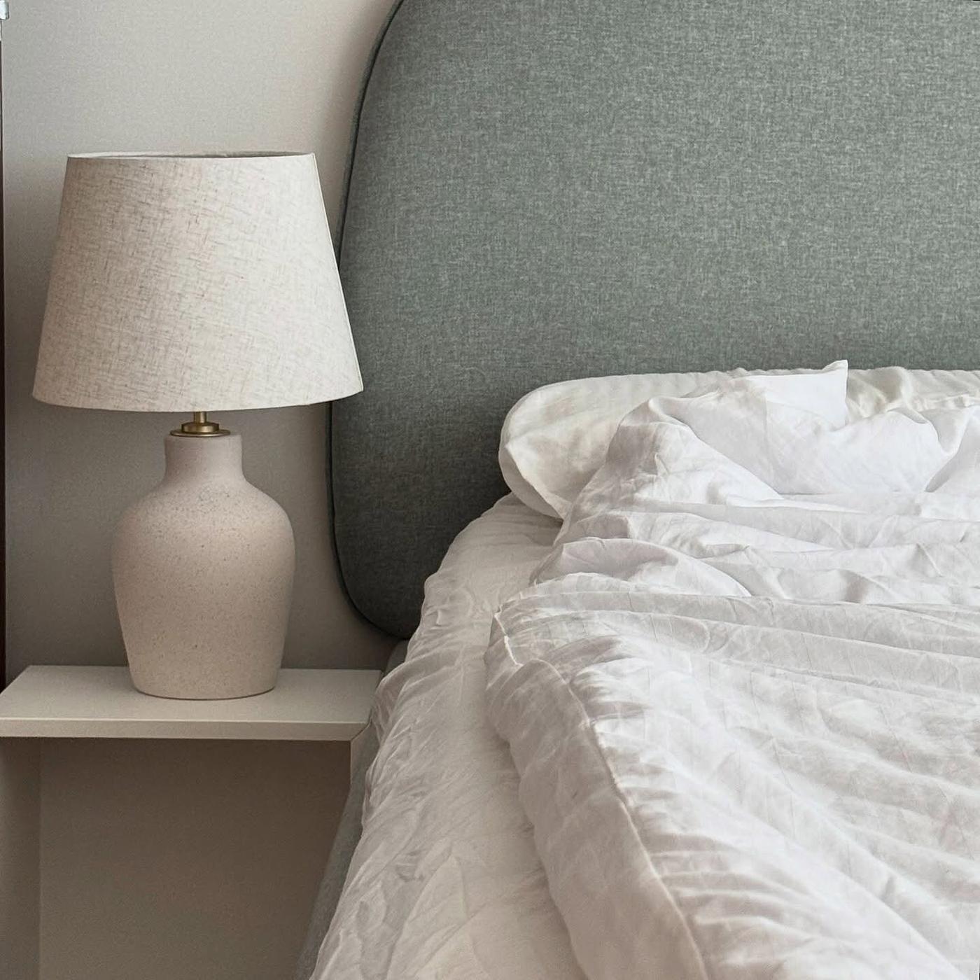 En sovrumsscen med en BLIDVÄDER bordslampa på ett vitt nattduksbord bredvid en säng med grå sänggavel och vita skrynkliga sängkläder.