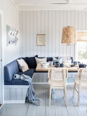 En spisestue med en hjørnebænk med blå hynder og lyse puder og ellers lyst interiør i et sommerhus. 