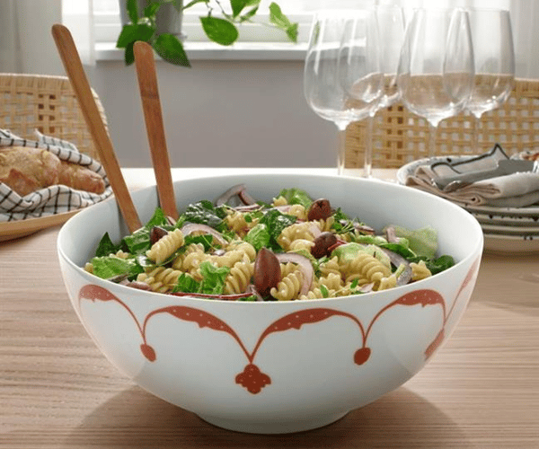 En stor vit GOKVÄLLÅ skål med rött mönster står på ett matbord. I skålen finns pastasallad och salladsbestick i trä. 