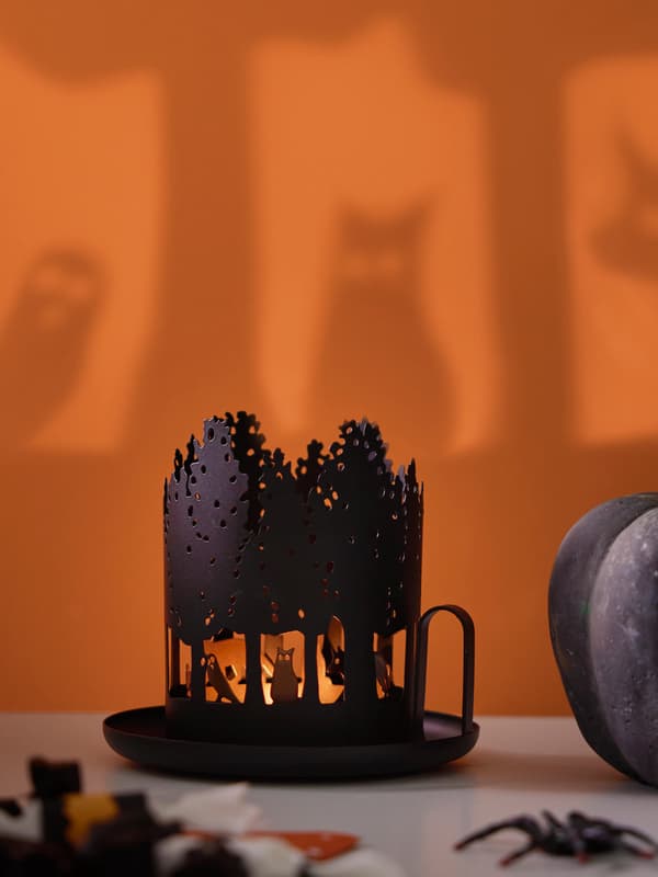 En svart ljuslykta med djurmönster är tänd och står på en vit bordsyta och kastar skuggor av djuren på en orange vägg.