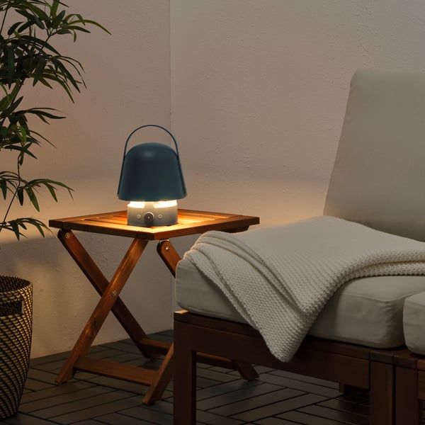 En tänd mörkblå lampa på ett träbord bredvid en vit soffa med en vit filt ovan