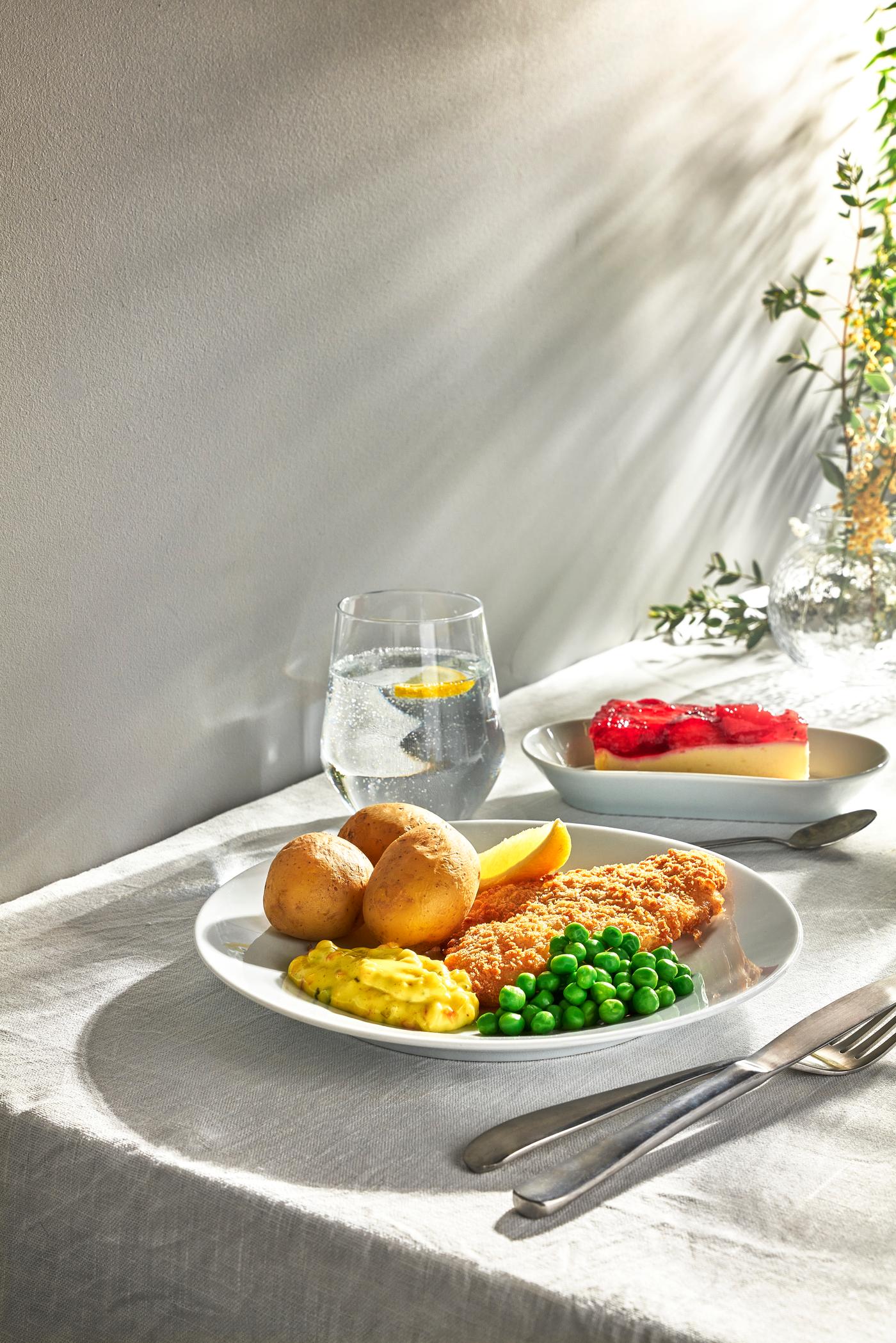 En tallrik med panerad fisk, potatis och ärtor på ett solbelyst bord bredvid ett glas vatten och en bit jordgubbscheesecake.