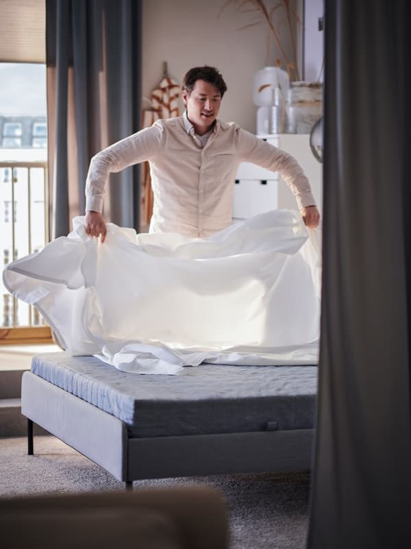 En una habitación iluminada, una persona coloca ropa de cama blanca sobre una estructura de cama SLATTUM tapizada en gris claro y un colchón.