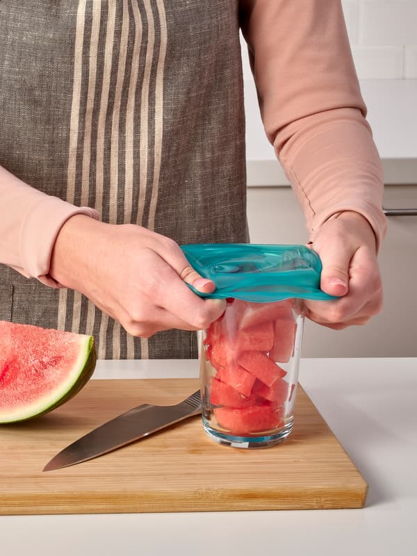 En una tabla de cortar de madera, una persona cubre un vaso de sandía picada con una cubierta de silicona reutilizable para alimentos ÖVERMÄTT.