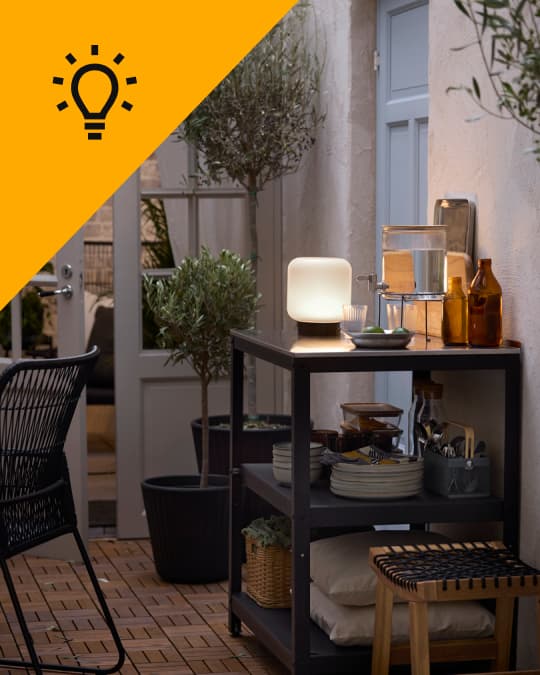 En una terraza hay un mueble con decoraciones y una lámpara inalámbrica de mesa encendida.