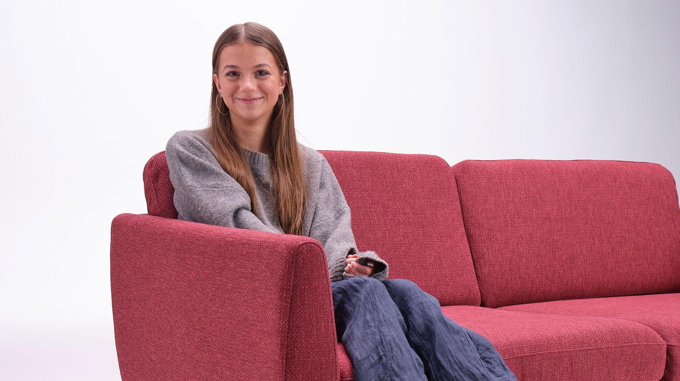 En ung kvinde med langt brunt hår sidder på en rød sofa i, hvad der ser ud til at være et IKEA showroom. Hun er klædt i en grå sweater og blå bukser og smiler blidt til kameraet med en afslappet kropsholdning, hvilket tyder på uendelige muligheder. Baggrunden er almindelig hvid.
