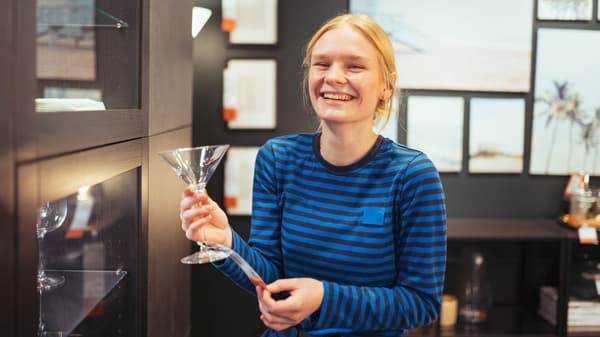 En ung person med lyst hår smiler og holder et martini-glas i en moderne, veloplyst butik. De er iført en blåstribet skjorte og står ved siden af glashylder med indrammede billeder synlige på væggen bag dem, der inkarnerer værdierne af en elev i IKEA.