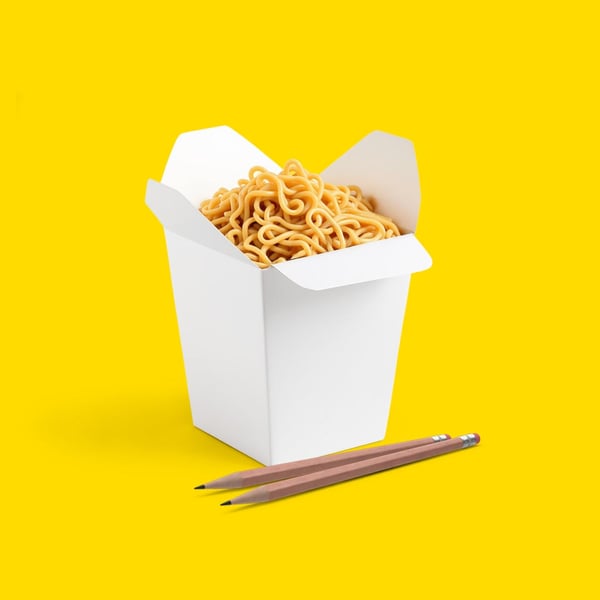 En vit takeaway-låda fylld med nudlar, placerad bredvid två blyertspennor, mot gul bakgrund.