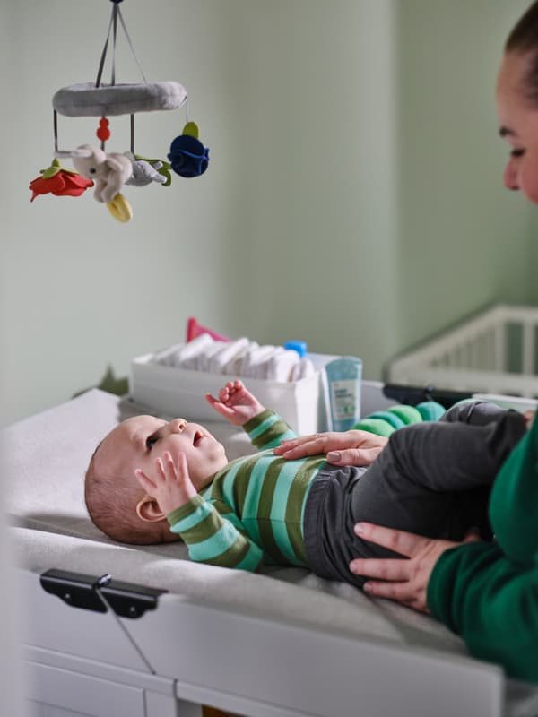 En voksen står over et lite, påkledd barn som ligger på et SUNDVIK stellebord og titter på en flerfarget GULLIGAST uro som henger fra taket.