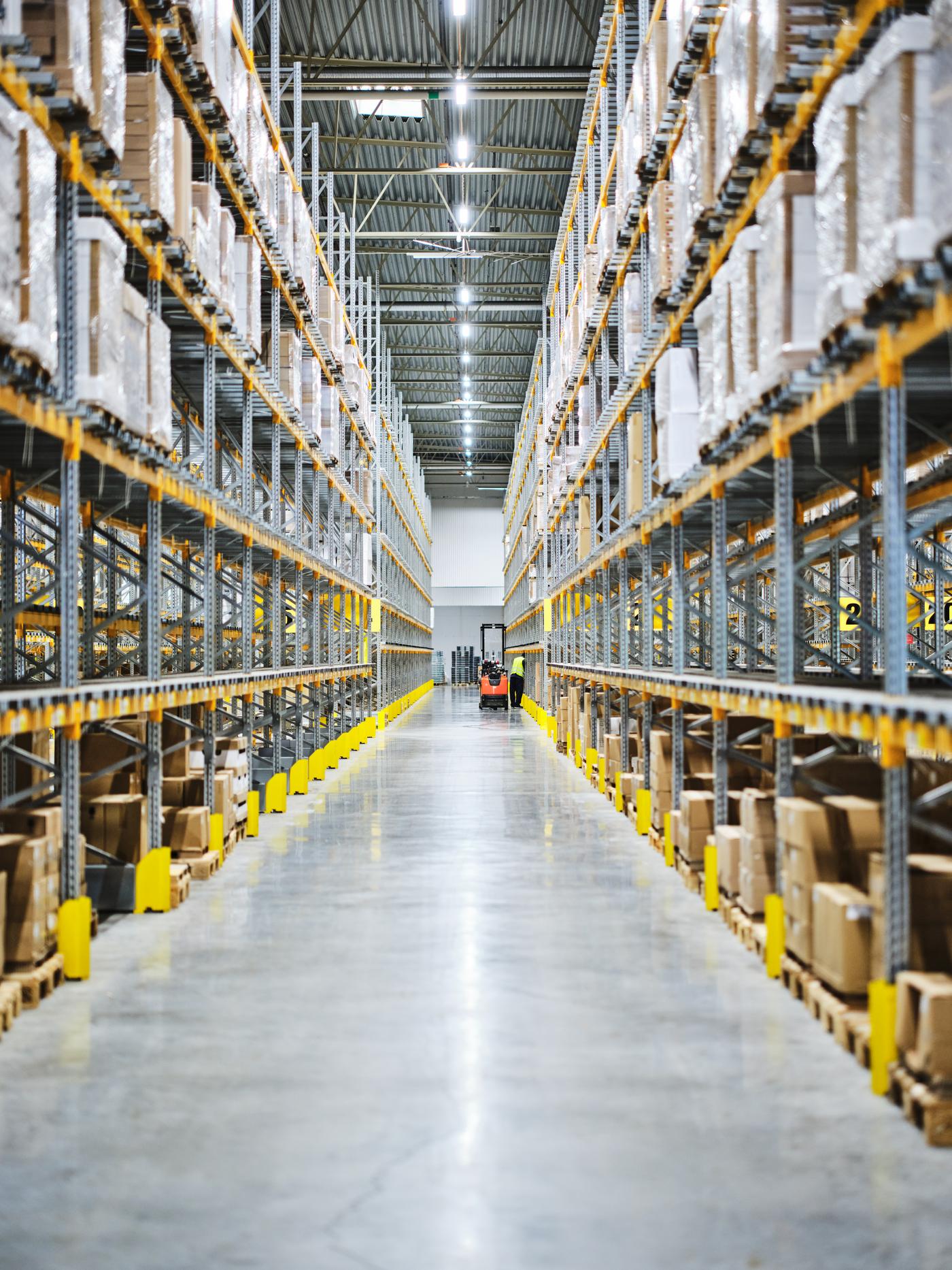 En vy av en lång gång i ett lager. Hyllorna är packade med varor från golv till tak på både högra och vänstra sidan. Längst bort i gången syns en medarbetare och en truck.