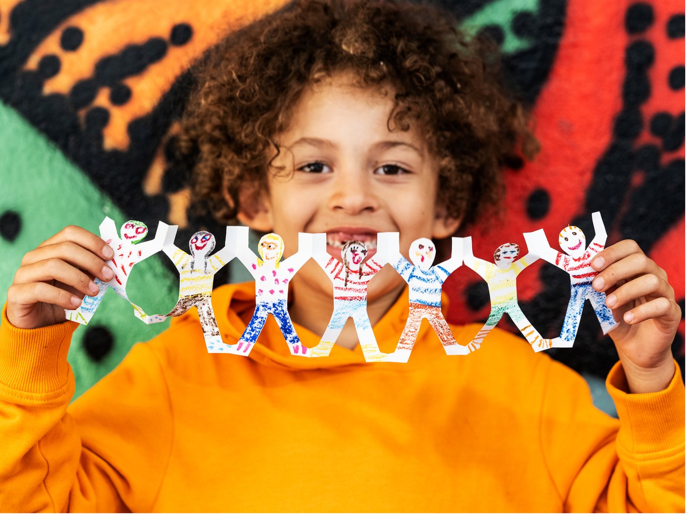 Enfant tenant une guirlande colorée en papier avec des personnages dessinés, devant un mur décoré de motifs graphiques vifs.