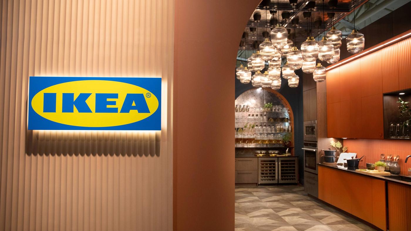 Enseigne IKEA sur un mur nervuré avec, en arrière-plan, une cuisine moderne équipée de suspensions en verre et d'armoires en bois.