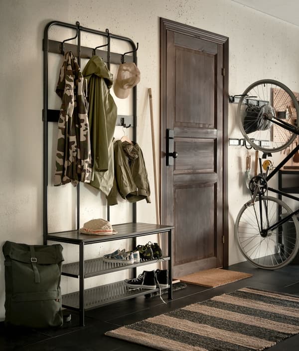 Entrada con un rack para ropa y zapatero, se ven varias prendas y calzado. Al otro lado de la puerta una bicicleta colgada de la pared.