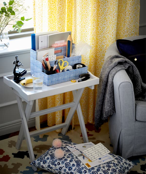 Entre une fenêtre et un fauteuil dans le salon, une table-plateau avec tout le nécessaire pour le travail scolaire, boisson et fruit y compris.