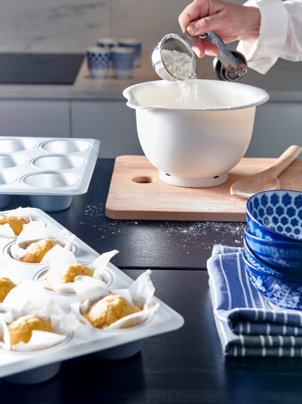 ภายในห้องครัวระหว่างการอบขนม: มือข้างหนึ่งเทแป้งลงในชาม ENTUSIASM/อันทูซิอาซึม ที่อยู่ถัดจากพิมพ์มัฟฟินที่อบเสร็จแล้ว