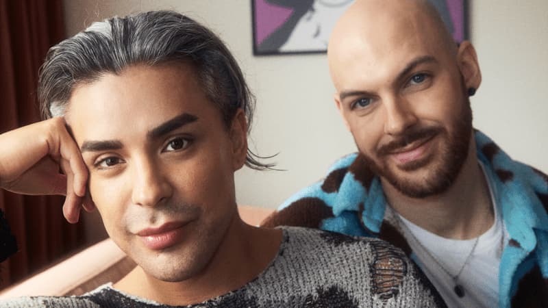 Envy Peru and her boyfriend sitting in their living room, a red curtain and a colorful painting in the background.