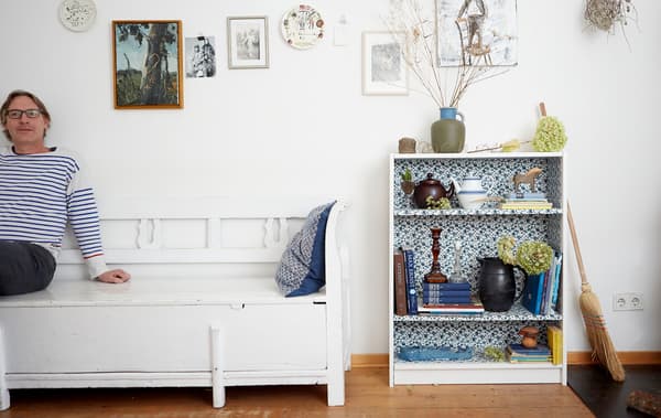 Erik assis sur un banc blanc contre un mur blanc, à côté d’une bibliothèque tapissée d’un tissu au motif floral et contenant des livres et des décorations.