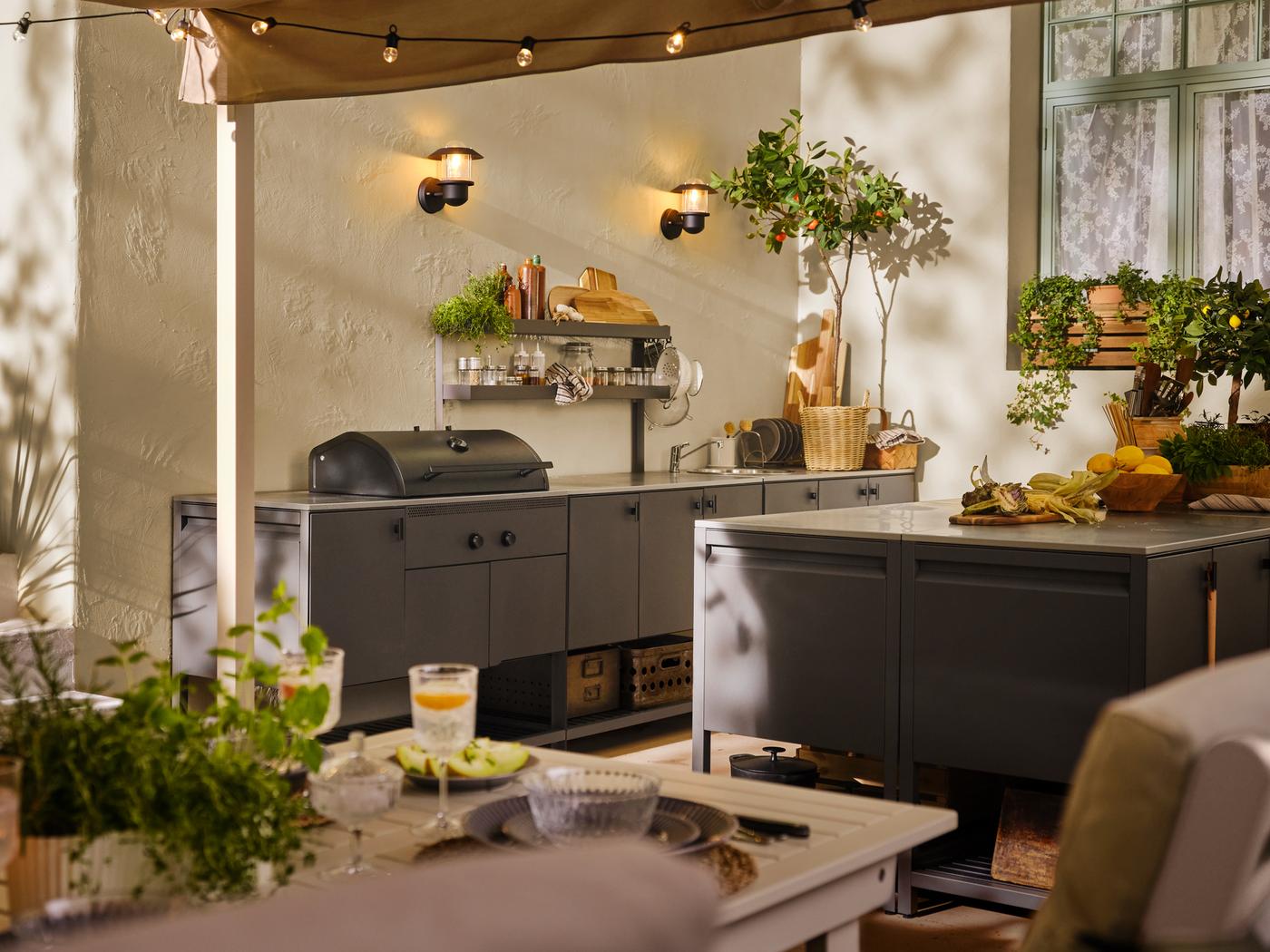 Es ist ein Aussenbereich mit einem BONDHOLMEN Tisch in Weiss/Beige und verschiedenen BÅTSKÄR Elementen in Dunkelgrau zu sehen, zu denen auch ein Kohlegrill gehört.