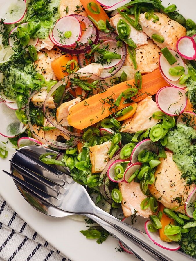 Es ist ein bunter Salat mit gegrilltem Lachs, Radieschen, Karotten, Zuckerschoten und frischen Kräutern zu sehen, der auf einem weißen Teller serviert wird.