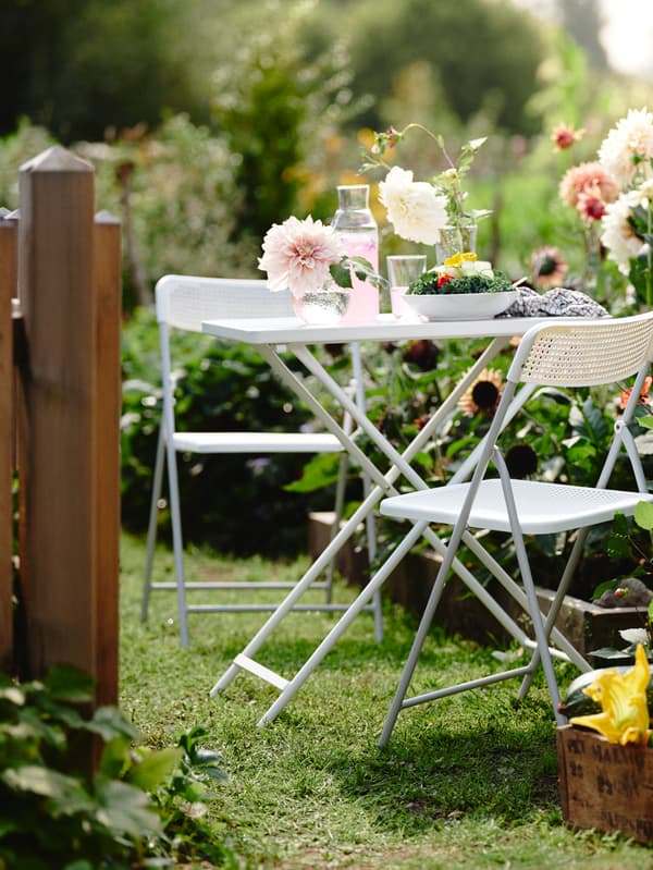 Es ist ein Garten mit vielen Blumen und zwei TORPARÖ Stühlen um einen TORPARÖ Tisch zu sehen. Auf dem Tisch stehen rosafarbene Getränke, Vasen und eine Schüssel Salat.