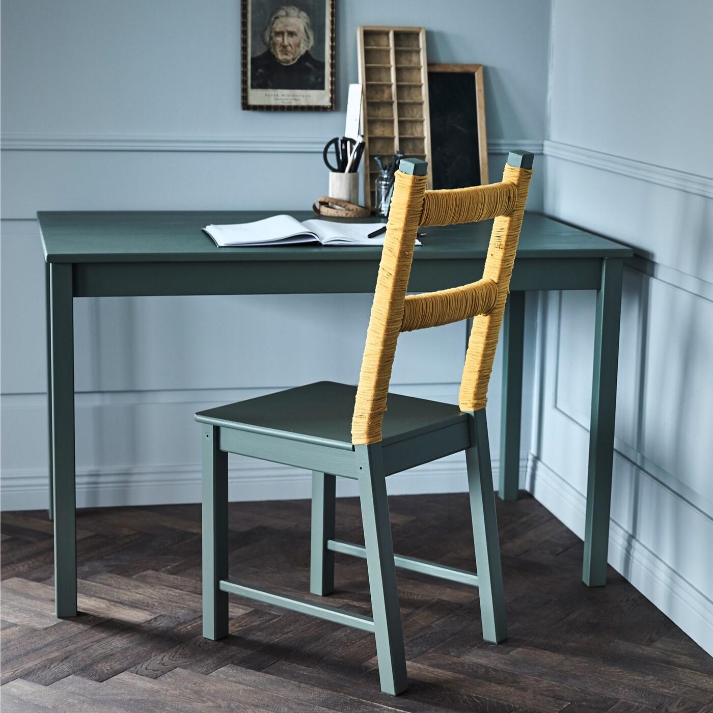 Es ist ein grüner Stuhl der an einem grünen Tisch steht worauf alte Bücher liegen und ein altes Bild an der Wand hängt.