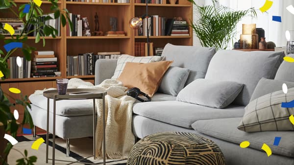 Es ist ein helles Wohnzimmer mit einem zentral platzierten SÖDERHAMN Sofa, TONSTAD Bücherregalen in Eichenfurnier, einem GLADOM Tabletttisch und großen Zimmerpflanzen zu sehen.