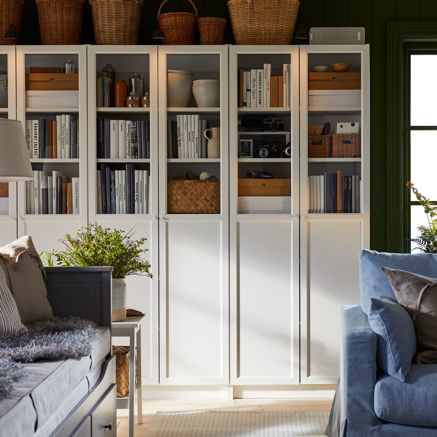 Es ist ein Raum mit BILLY Bücherregalen mit Paneel-/Vitrinentüren hinter einem blauen Sofa und einem weißen Couchtisch zu sehen. In den Bücherregalen befinden sich Bücher und Geschirr.