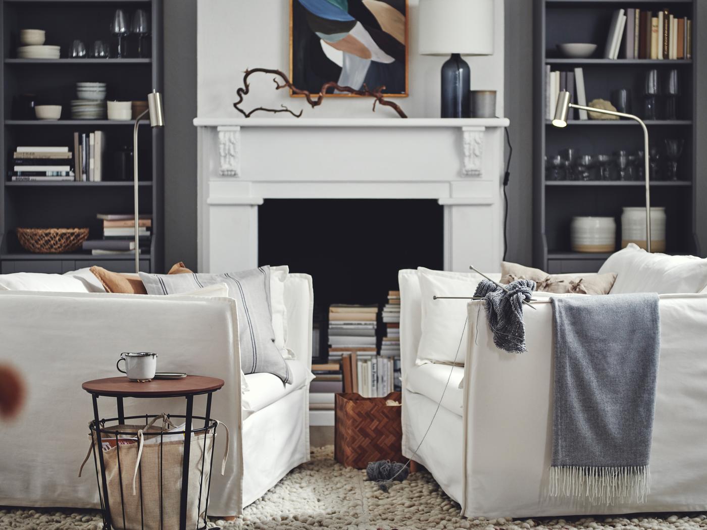 Es ist ein Wohnzimmer zu sehen, in dem sich zwei weiße BACKSÄLEN 1,5-Sessel gegenüberstehen. Im Hintergrund befinden sich graue HAUGA Hochschränke rechts und links eines Kamins.