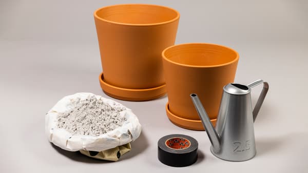 Es sind 2 Tontöpfe in unterschiedlichen Größen, eine Gieskanne, Sand und ein breites Klebeband zu sehen. 