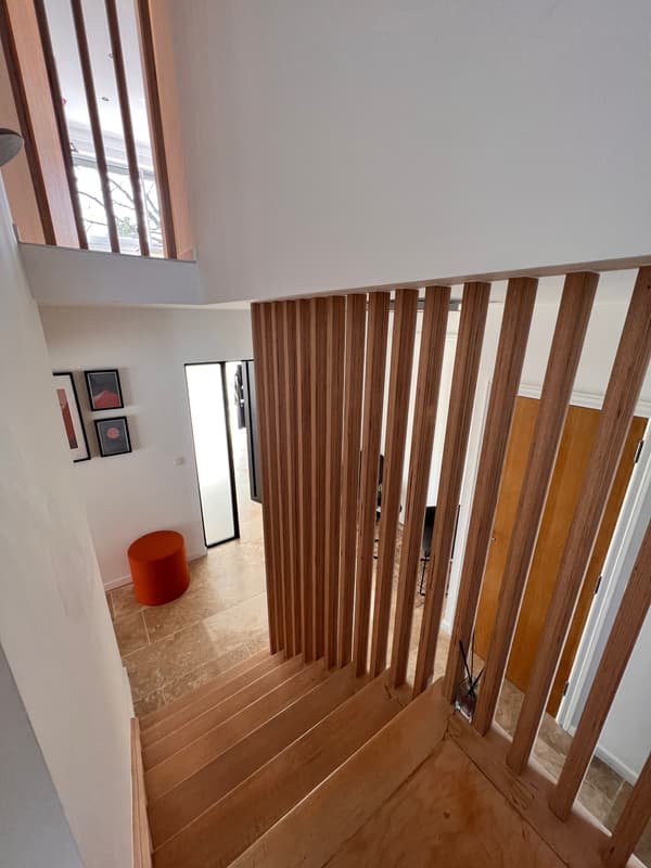 Escalier intérieur en bois avec garde-corps à lattes verticales, menant à un hall lumineux avec murs blancs et tabouret rouge.