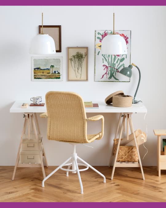 Escritorio blanco con silla de fibra trenzada, lámparas colgantes y cuadros decorativos en la pared.