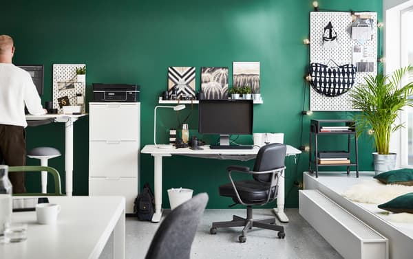 Escritorios para trabajar sentado y de pie BEKANT blancos en una oficina con las paredes verdes con sillas giratorias ALEFJÄLL negras y un taburete NILSERIK.