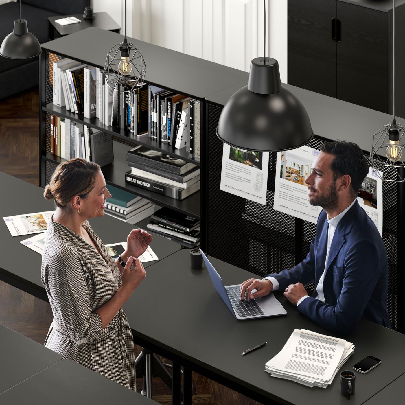 Espace bureau où une femme parle à un homme vêtu d’un costume assis à une table TOMMARYD avec son ordinateur portable.