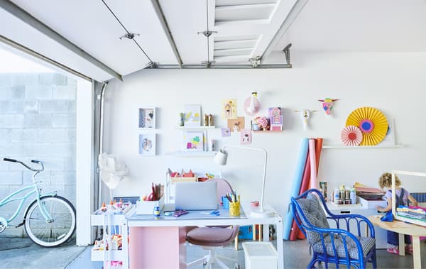 Espace de travail coloré avec bureau, desserte, chaises et présentoirs muraux, dans un garage avec la porte à enroulement ouverte.