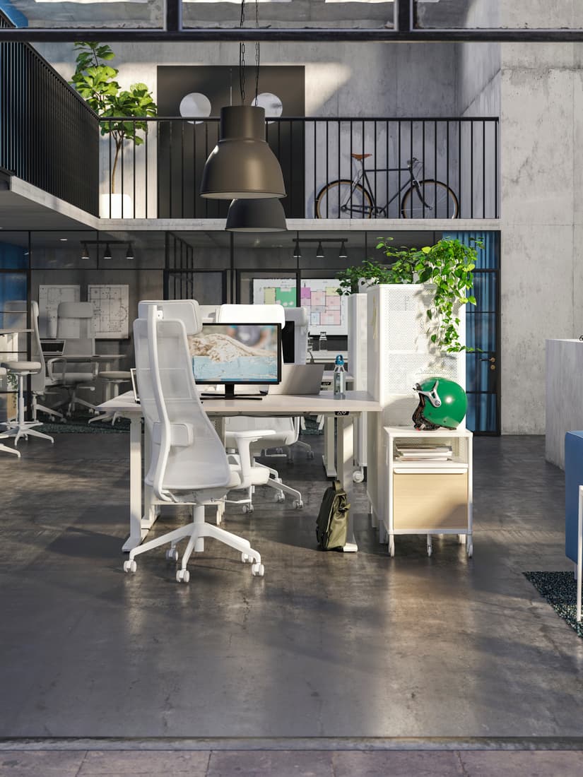 Espacio de trabajo con escritorio RODULF y silla de escritorio RODULF, al lado derecho del escritorio una cajonera con una planta encima. En el fondo se ve una terraza interior con una biclicleta en el segundo piso, en el primer piso se ven puestos de trabajo.
