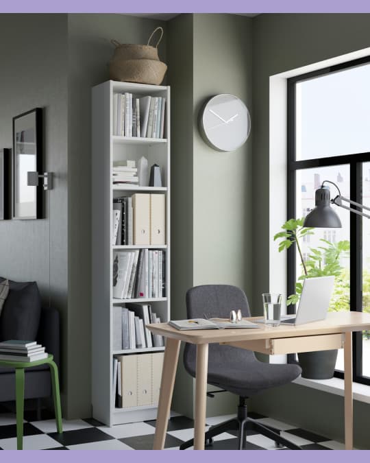 Espacio de trabajo con una biblioteca alta y delgada color blanco, llena de libros y pastas; al lado, un escritorio de madera clara junto a una ventana grande.