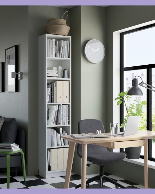 Espacio de trabajo con una biblioteca alta y delgada color blanco, llena de libros y pastas; al lado, un escritorio de madera clara junto a una ventana grande.