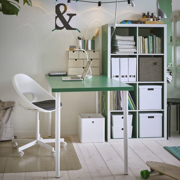 Espaço de estudo com uma mesa com o tampo verde e pernas em branco, uma cadeira giratória em branco, um candeeiro de secretária em branco e um protetor de chão transparente.