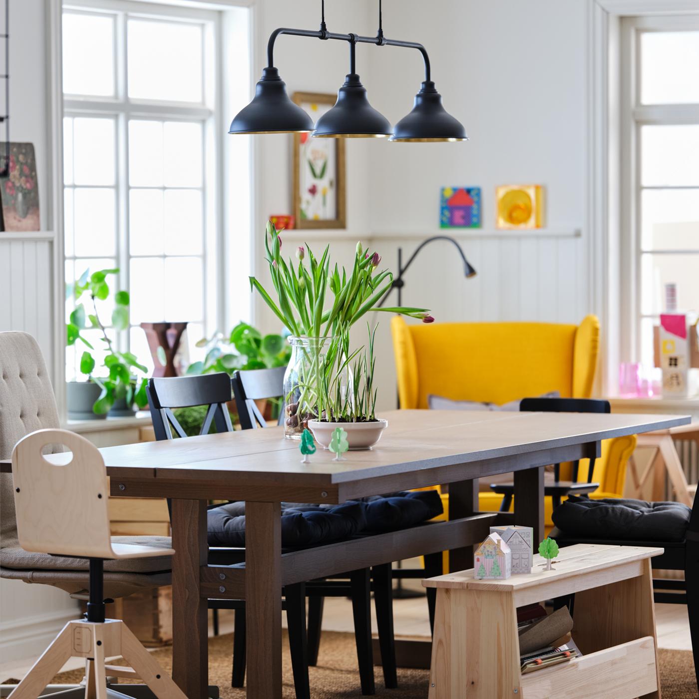 Espaço de refeição com uma mesa, cadeiras e candeeiros numa divisão grande, iluminada e arejada.
