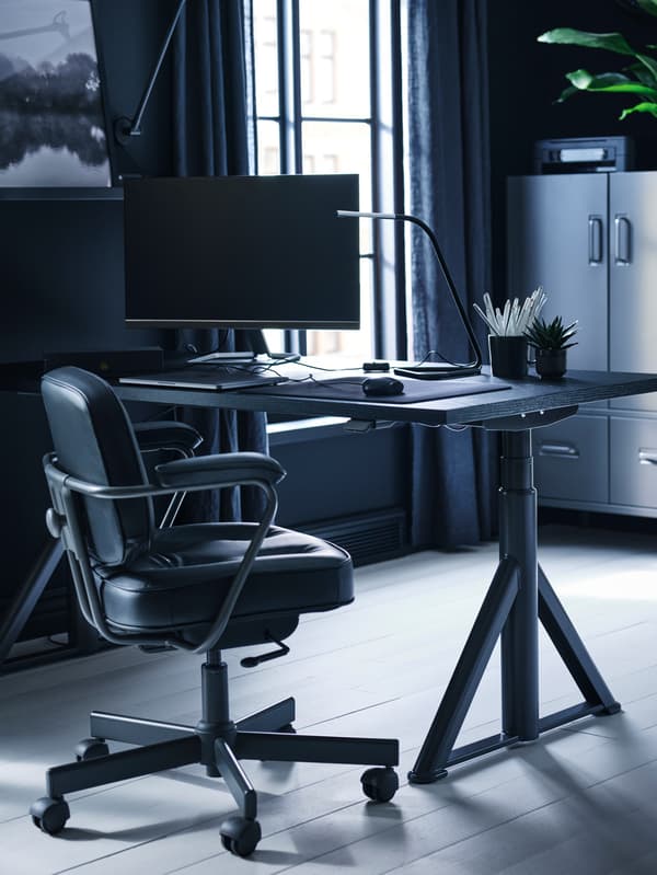 Espaço de trabalho em casa com secretária sentado/em pé IDÅSEN em preto/cinzento escuro junto a uma janela, com um quadro numa moldura em preto numa das paredes.