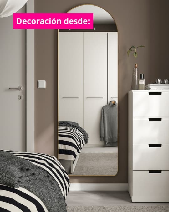 Espejo de cuerpo completo con marco dorado apoyado en un dormitorio, reflejando muebles blancos y ropa ordenada. Texto en imagen dice: “Decoración desde:”.