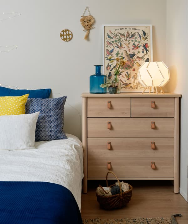 Esquina dunha cama con roupa azul e branca xunto a unha cómoda nun cuarto branco.